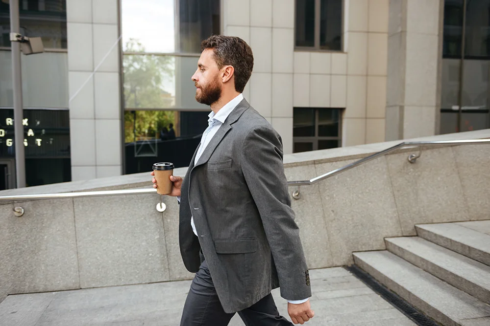Man in gray suit holding takeaway coffee and walking along street